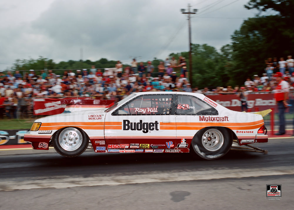 Roy Hill Topaz Race car on a track with 'Budget Motorcraft' branding in front of a crowd.